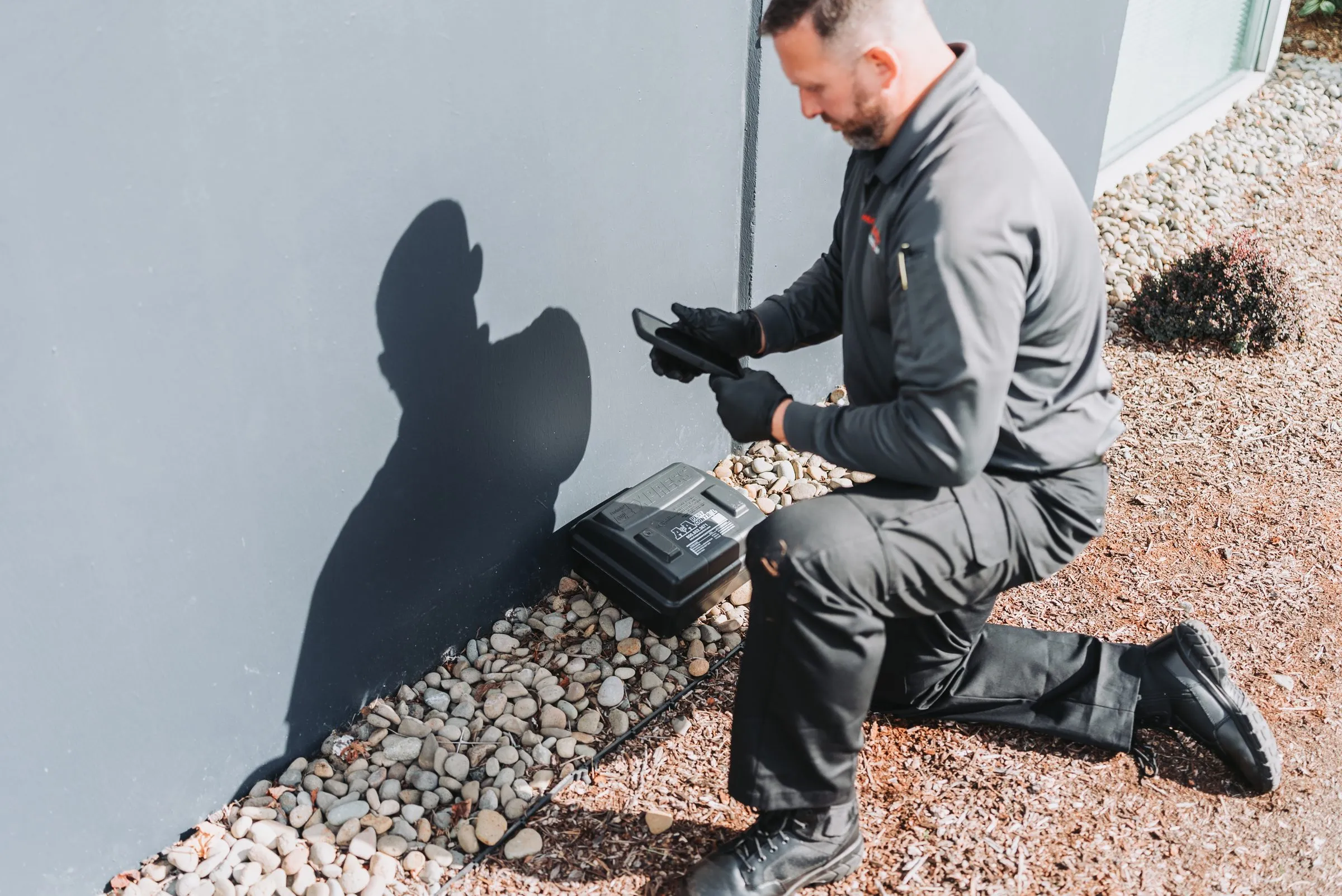 An A&A technician servicing an exterior rodent station along a building's foundation