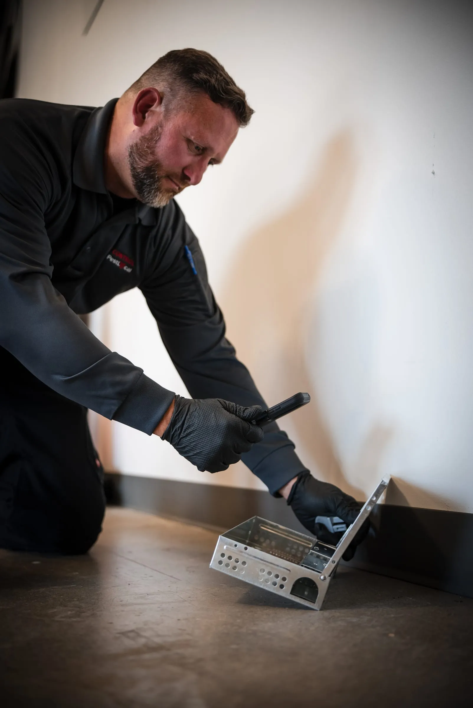An A&A technician checking a multiple-catch rodent trap along an interior wall with a flashlight