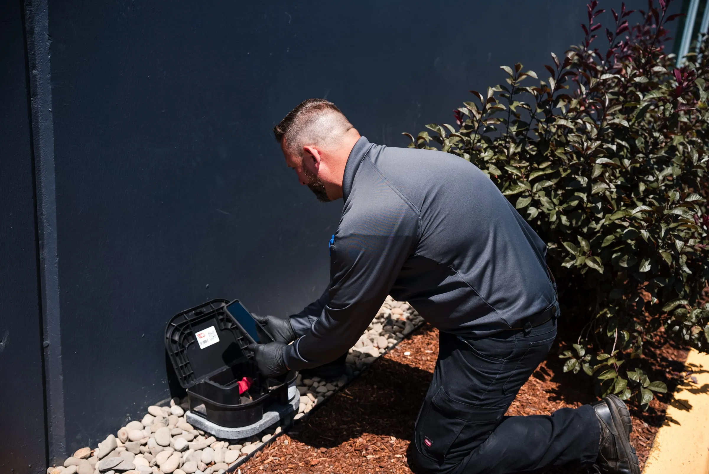 An A&A technician kneeling to open an exterior rodent bait station beside a building