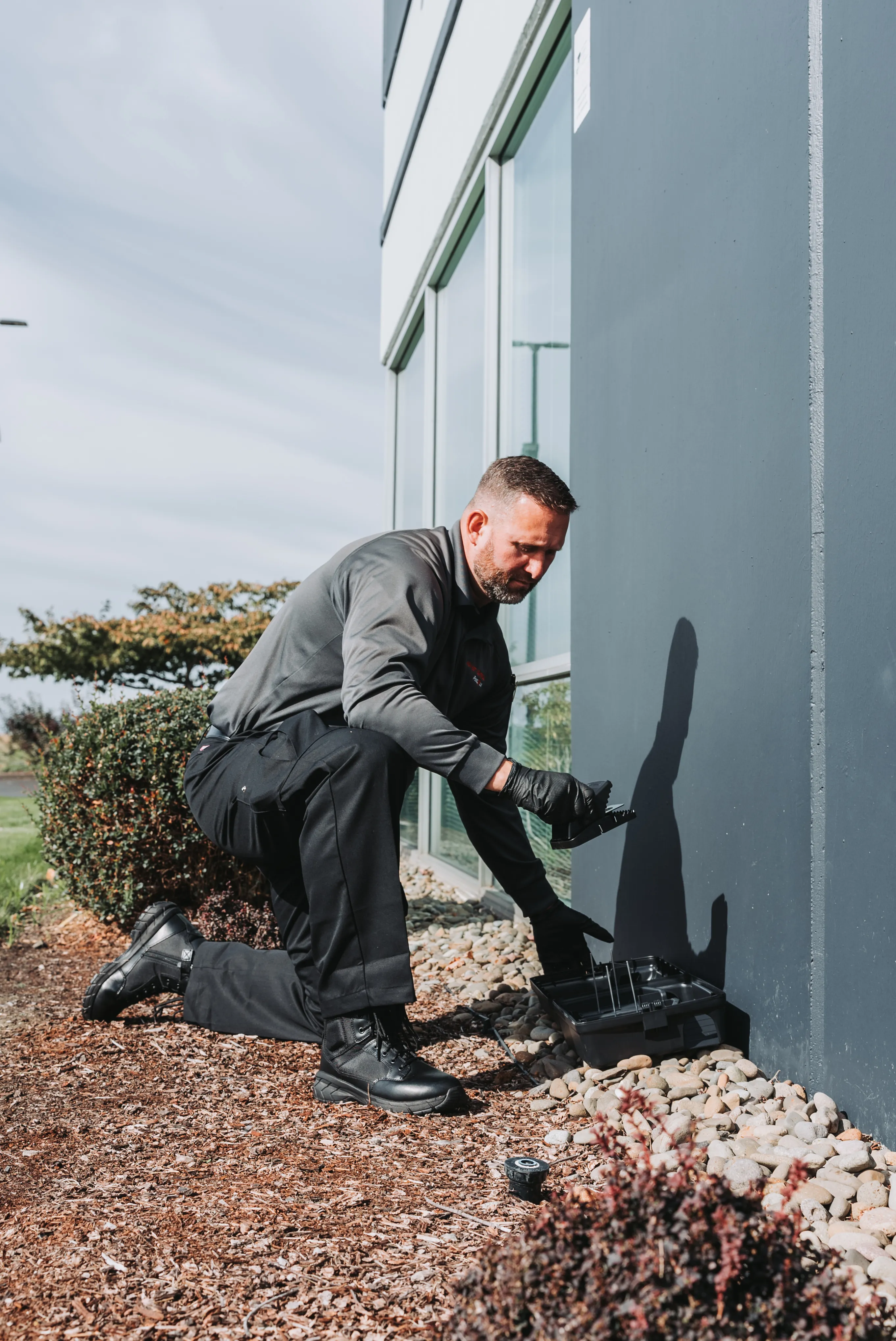 An A&A Pest Control technician kneeling at the base of a building to service an exterior rodent bait station