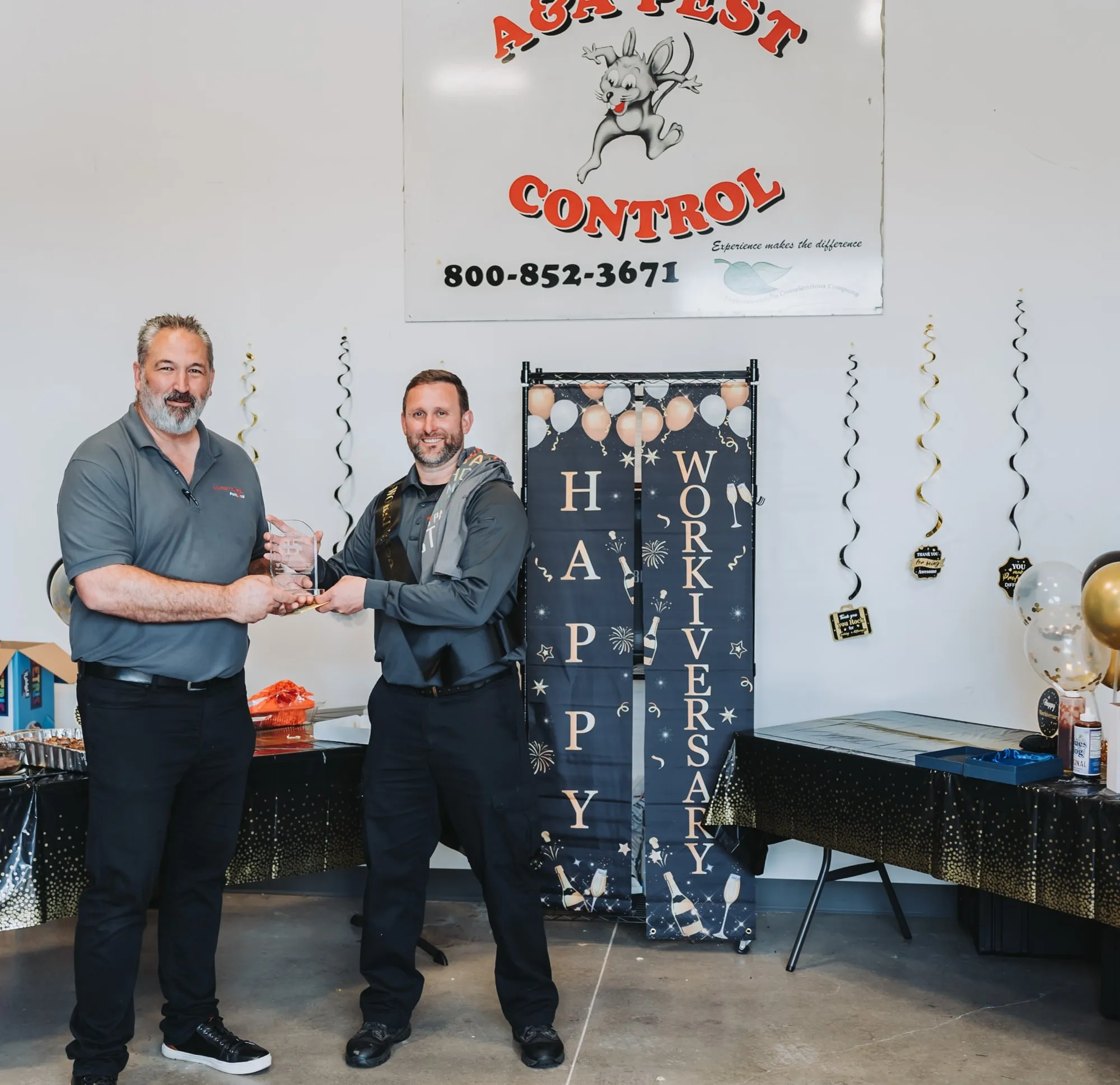 Steve Wittig presenting a team member with an award at an A&A work anniversary celebration, under a Happy Workiversary banner